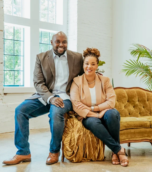 A casually dressed man and woman sitting together on stylish furniture in a bright, modern room with large windows and indoor plants.